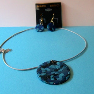 May include: A jewelry set featuring a necklace and matching earrings. The necklace has a round pendant with a blue and teal abstract design. The earrings are small, round, and have a similar pattern. The set is displayed on a light blue surface.