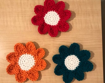 May include: Three crocheted flower coasters in red, orange, and teal. Each coaster has a white center.