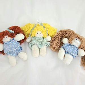 May include: Three handmade fabric dolls with yarn hair and cloth bodies. The left doll has auburn hair and a blue floral romper. The center doll has yellow hair and a green floral romper. The right doll has light brown hair and a blue floral romper.