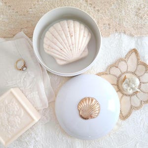 May include: A white round container with a gold seashell emblem on the lid, containing a pale pink seashell-shaped soap. Beside it sits a rectangular soap bar with floral embossing, a gold ring, and a small white decorative angel figurine.
