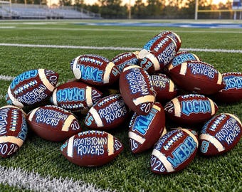 May include: A pile of brown leather footballs with white laces, each customized with blue painted text reading Rootstown Rovers, individual names, and the year 2026. The footballs feature white stripes and are arranged on a green turf field.