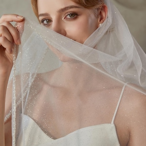 May include: A sheer white wedding veil with a delicate beaded edge and scattered glitter. The veil is draped over a white spaghetti strap dress. The image is a close-up of the bride's face, with the veil partially obscuring her features.