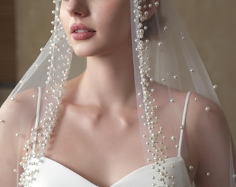 May include: A bridal veil and headband adorned with numerous small, round pearls. The sheer veil cascades over a white dress with spaghetti straps. The pearls are clustered along the edges of the veil and headband.