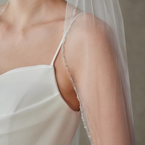 Può includere: Un velo da sposa bianco trasparente con un delicato bordo di perline. Il velo è drappeggiato su un abito di raso bianco, creando un look morbido ed elegante. Le perline sono piccole e scintillanti, aggiungendo un tocco di glamour al design.