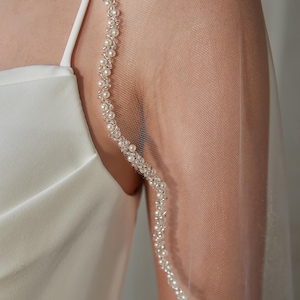 May include: A sheer white bridal veil with a delicate pearl trim. The veil is draped over the shoulder, showcasing the intricate pearl detailing. The dress is a simple white satin. The overall aesthetic is elegant and classic.