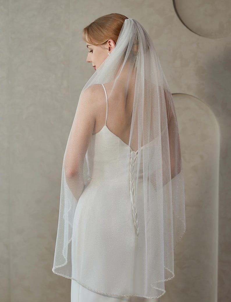 May include: A bride wearing a white wedding dress and a sheer, sparkling veil. The veil is trimmed with a delicate beaded edge. The dress has a low back and spaghetti straps.