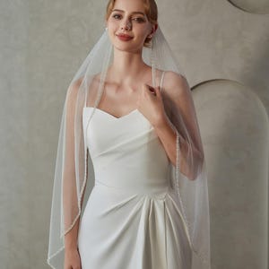 May include: A bride wearing a white wedding dress with a sweetheart neckline and a high slit. She is wearing a sheer veil with a beaded trim and scattered embellishments. The veil is draped over her shoulders.