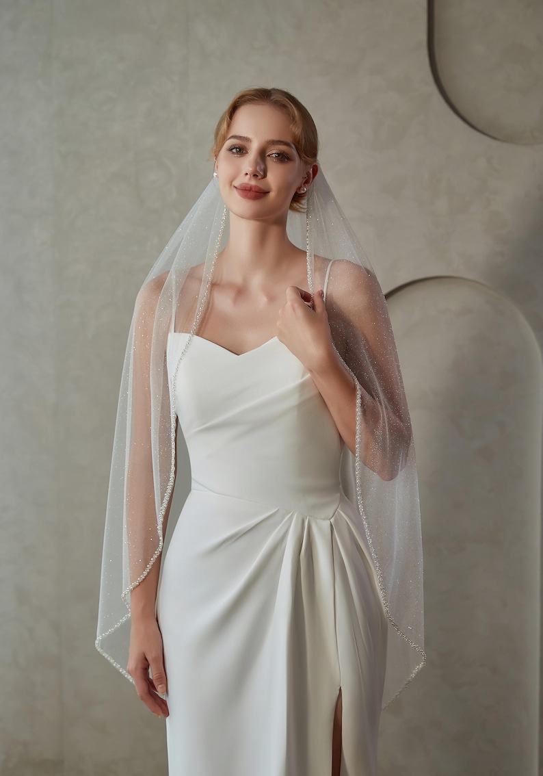 May include: A bride wearing a white wedding dress with a sweetheart neckline and a high slit. She is wearing a sheer veil with a beaded trim and scattered embellishments. The veil is draped over her shoulders.