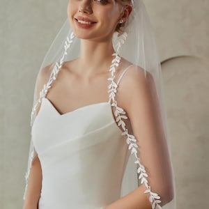 Peut inclure: Une robe de mariée blanche avec un décolleté en cœur et un voile transparent. Le voile est orné de broderies blanches délicates en forme de feuilles sur les bords. La mariée sourit, les cheveux attachés.