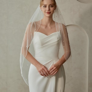 May include: A white wedding dress with a sweetheart neckline and a slit. A sheer veil is adorned with small pearls along the edges. The bride is wearing pearl earrings.