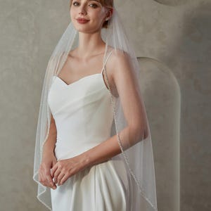May include: A bride wearing a white wedding dress with a sweetheart neckline and a high slit. She is also wearing a sheer veil with a beaded edge. The veil is draped over her head and shoulders.