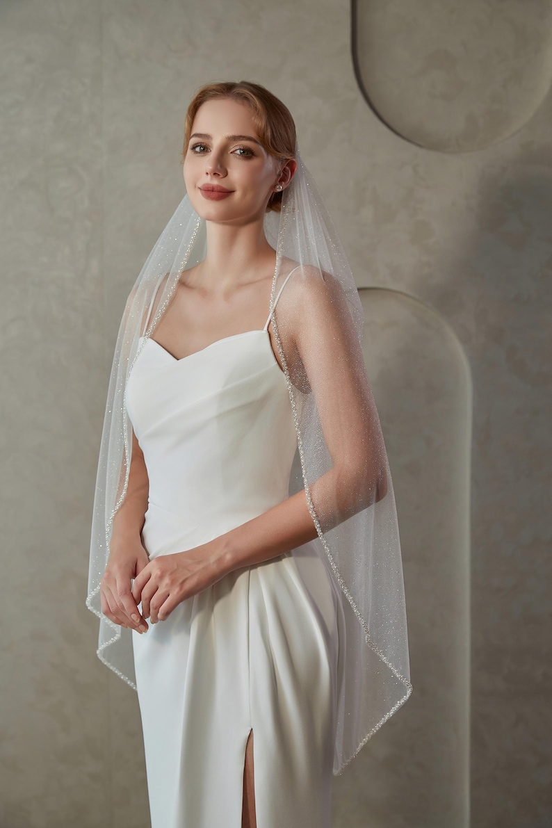 May include: A bride wearing a white wedding dress with a sweetheart neckline and a high slit. She is also wearing a sheer veil with a beaded edge. The veil is draped over her head and shoulders.