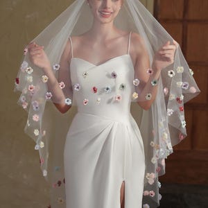 Op de afbeelding: Een witte trouwjurk met een hoge split en spaghettibandjes. De jurk is versierd met kleurrijke bloemaccenten. Een doorschijnende sluier, eveneens versierd met bloemen, wordt omhoog gehouden, wat een romantische en elegante uitstraling creëert.