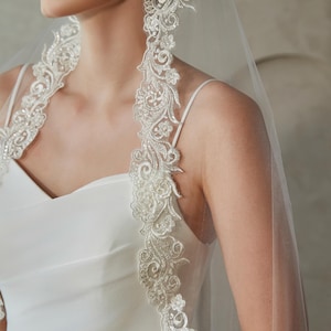 Op de afbeelding: Een witte bruidssluier met ingewikkelde bloemenkanten details langs de randen. De sluier is doorschijnend en het kant is versierd met kleine, sprankelende versieringen. De bruid draagt een witte jurk.