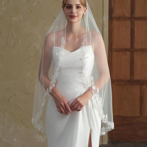 May include: A bride wearing a white wedding dress with a high slit and a sheer veil. The veil is trimmed with delicate floral lace. The dress has a sweetheart neckline and spaghetti straps.