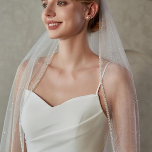 May include: A bride wearing a white wedding dress and a sheer veil. The veil is adorned with small, sparkling embellishments and a delicate beaded trim. The dress features a simple, elegant design with spaghetti straps.