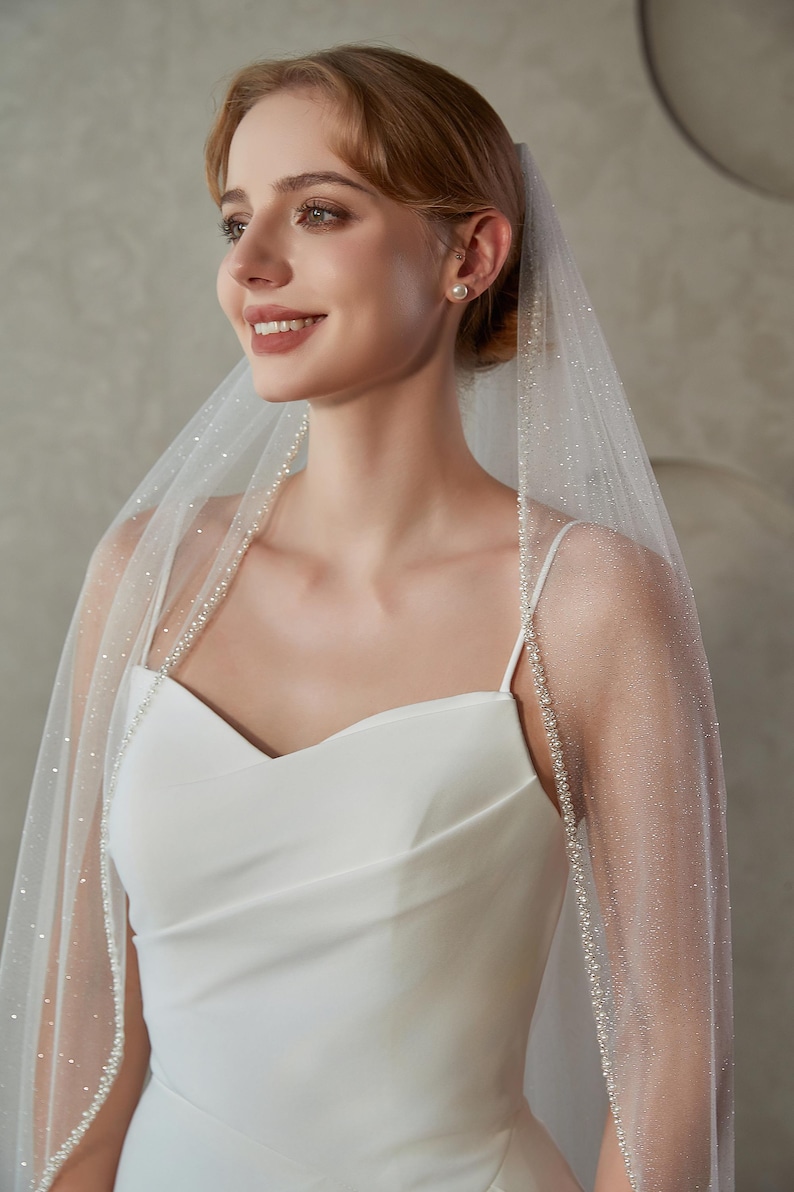 May include: A bride wearing a white wedding dress and a sheer veil. The veil is adorned with small, sparkling embellishments and a delicate beaded trim. The dress features a simple, elegant design with spaghetti straps.