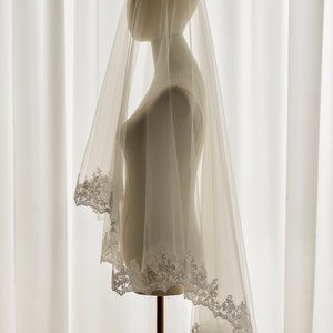 May include: A white bridal veil with a delicate lace trim. The veil is draped over a mannequin and is made of soft, sheer fabric.