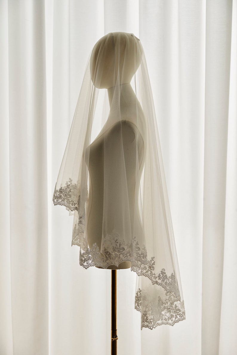 May include: A white bridal veil with a delicate lace trim. The veil is draped over a mannequin and is made of soft, sheer fabric.