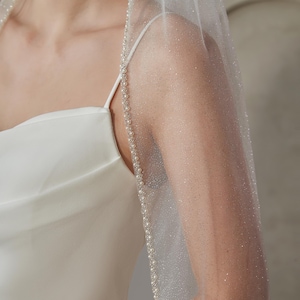 May include: A sheer, sparkling wedding veil with a delicate pearl trim. The veil is draped over a white satin dress, creating a soft, ethereal look. The veil's edge is adorned with small, evenly spaced pearls.