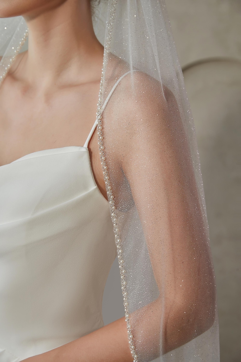 May include: A sheer, sparkling wedding veil with a delicate pearl trim. The veil is draped over a white satin dress, creating a soft, ethereal look. The veil's edge is adorned with small, evenly spaced pearls.