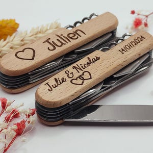 Op de afbeelding: Twee houten multitools met diverse gereedschappen. Eén is gegraveerd met "Julien" en een hart, de andere met "Julie & Nicolas", twee harten en "14/07/2024". De tools hebben een kurkentrekker, mes, vork en lepel.