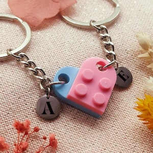 Peut inclure: Un porte-clés en forme de cœur divisé, composé de briques en plastique roses et bleues, avec des porte-clés et des maillons de chaîne argentés. Chaque moitié porte un charme circulaire avec les lettres "A" et "E". Fond en tissu rose clair avec des fleurs séchées.