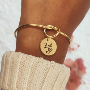 Könnte beinhalten: Goldfarbenes Armreif-Armband mit Knoten-Design und rundem Anhänger. Der Anhänger ist mit dem Namen "Zoé" und zwei Herzen graviert. Das Armband wird am Handgelenk getragen, mit einem cremefarbenen Pullover im Hintergrund.