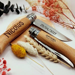 Pode incluir: Duas facas Opinel com cabo de madeira, uma gravada com "Nicolas" e a outra com "29/04/2021" e "Opinel Savoie France". As facas s&atilde;o exibidas com flores secas e espigas de trigo, sugerindo um presente personalizado.