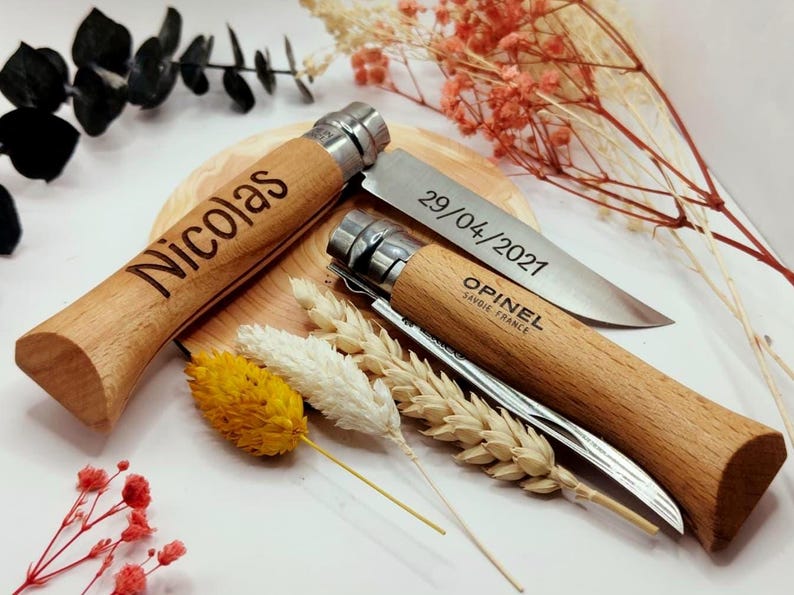 Pode incluir: Duas facas Opinel com cabo de madeira, uma gravada com "Nicolas" e a outra com "29/04/2021" e "Opinel Savoie France". As facas s&atilde;o exibidas com flores secas e espigas de trigo, sugerindo um presente personalizado.