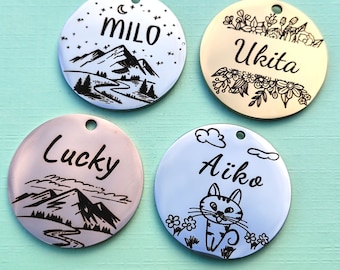 May include: Four round pet tags with engraved designs. One tag is white with "Milo" and a mountain scene. Another is gold with "Ukita" and floral accents. A third is pink with "Lucky" and a mountain scene. The last is blue with "Aiko" and a cat.