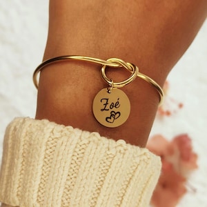Könnte beinhalten: Goldfarbenes Armreif-Armband mit Knoten-Design und rundem Anhänger. Der Anhänger ist mit dem Namen "Zoé" und zwei Herzsymbolen graviert. Das Armband wird am Handgelenk getragen, mit einem cremefarbenen Pullover im Hintergrund.