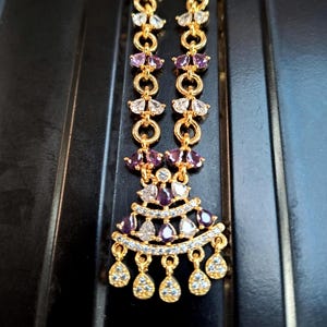 May include: Gold-toned necklace with a symmetrical design. The necklace features a series of small, round links connecting sections adorned with purple and clear gemstones. The pendant has a fan-like shape with teardrop-shaped dangles.