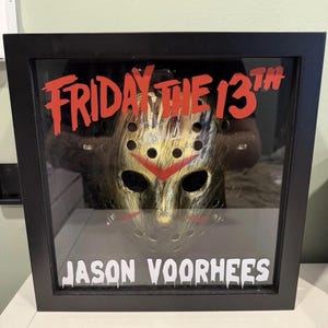 May include: A framed display featuring a hockey mask with a red chevron and gold accents. The text "FRIDAY THE 13TH" is in red above the mask, and "JASON VOORHEES" is in white below.