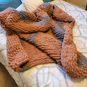 Può includere: Una coperta in maglia grossa in tonalità di marrone e grigio. La coperta è realizzata in filato spesso e ha una texture morbida e accogliente.
