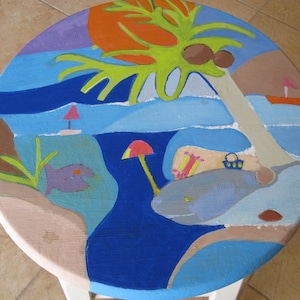 May include: A round wooden stool painted with a whimsical beach scene. The stool features a blue ocean, a palm tree with coconuts, a pink mushroom, a purple fish, and a light blue whale.