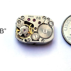 Peut inclure: Un petit mouvement de montre de couleur argent avec des engrenages dor&eacute;s et un rubis rouge. Le mouvement est &agrave; peu pr&egrave;s de la taille d'une pi&egrave;ce de dix cents.