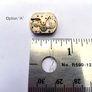 Peut inclure: Gros plan d'un petit mouvement de montre de couleur argent avec des engrenages visibles. Le mouvement mesure environ 2,5 cm de diam&egrave;tre et est pos&eacute; sur une r&egrave;gle avec le chiffre "1" visible.