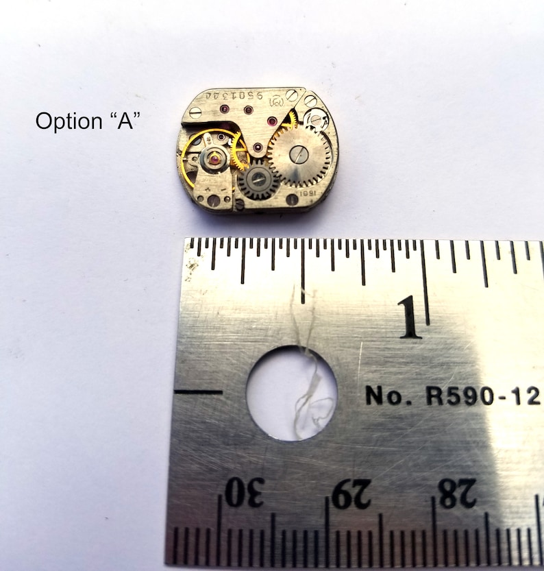 Peut inclure: Gros plan d'un petit mouvement de montre de couleur argent avec des engrenages visibles. Le mouvement mesure environ 2,5 cm de diam&egrave;tre et est pos&eacute; sur une r&egrave;gle avec le chiffre "1" visible.