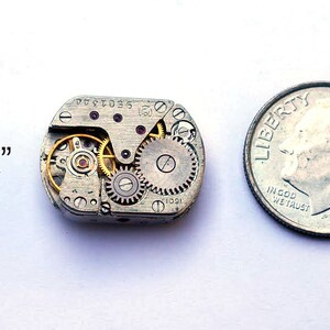 Peut inclure: Un petit mouvement de montre argent&eacute; avec des engrenages visibles et un rubis rouge. Le mouvement est &agrave; peu pr&egrave;s de la taille d'une pi&egrave;ce de dix cents.