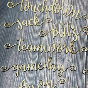 May include: A set of seven wooden cutouts with words related to American football, including "touchdown", "sack", "blitz", "teamwork", "gameday", "down", and "set".