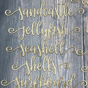 May include: Five wooden cutout words with a cursive font and a decorative swirl at the end of each word. The words are "sandcastle", "jellyfish", "seashell", "shells", and "surfboard".