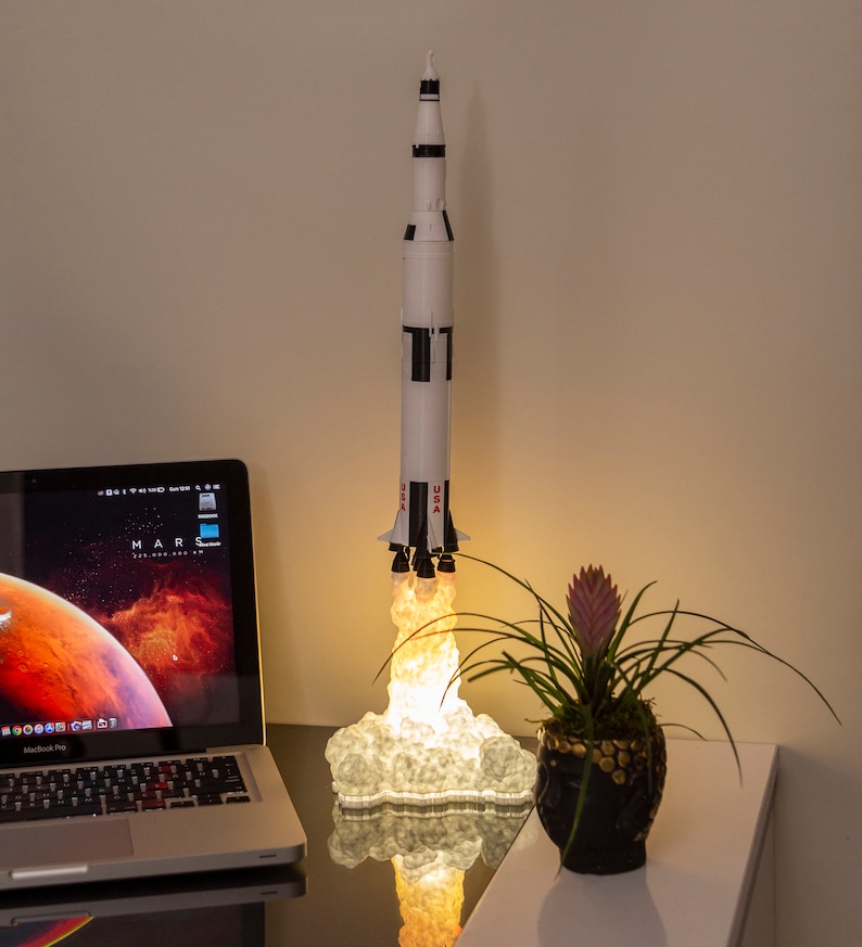 Może przedstawiać: Biało-czarny model rakiety Saturn V z rozgrzaną pomarańczową podstawą, przypominającą ognisty start. Rakieta stoi na biurku z laptopem i doniczką z rośliną.