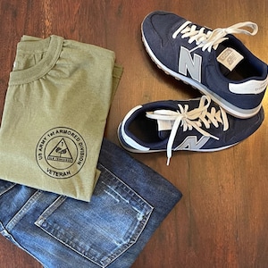 May include: A green t-shirt with a black circle logo that says "US Army 1st Armored Division Veteran". The t-shirt is folded and placed on top of a pair of blue jeans. A pair of dark blue sneakers with white laces and a white "N" logo are on a wooden surface.