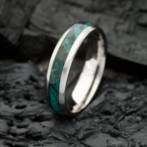 May include: A polished silver-toned metal ring featuring a central inlay of swirling teal and dark blue resin. The band has a smooth, reflective finish and a rounded profile, showcasing a distinct contrast between the metallic edges and the colorful center.