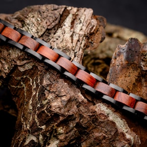 May include: A link bracelet featuring alternating segments of polished black metal and smooth reddish-brown wood. The design consists of rectangular wooden inserts set within black metal frames, creating a continuous chain pattern with a visible grain.