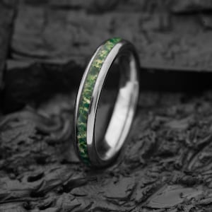 May include: A polished silver-toned metal ring featuring a central inlay of crushed green and gold mineral fragments. The band has a smooth, rounded exterior profile and a reflective finish that contrasts with the textured, earthy inlay.