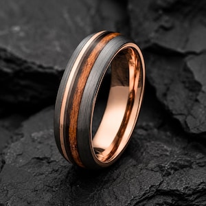 May include: A ring featuring a brushed dark gray metal band with a central inlay of brown wood and thin rose gold-colored accents. The interior of the band has a smooth, polished rose gold-colored finish, creating a contrast with the exterior.