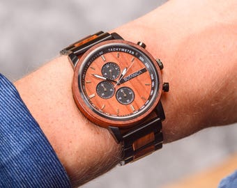 May include: A brown and black wooden wristwatch with a chronograph face. The watch face has a black background with white markings and the word "BOTANICA" printed in white. The watch has a black band with brown wood accents.