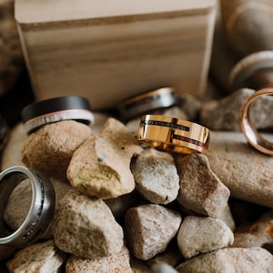 May include: A collection of rings in various styles and materials, displayed on a bed of small rocks. Rings include a gold band with black and red inlays, a black band with a wood-like inlay, and a silver ring with black accents.